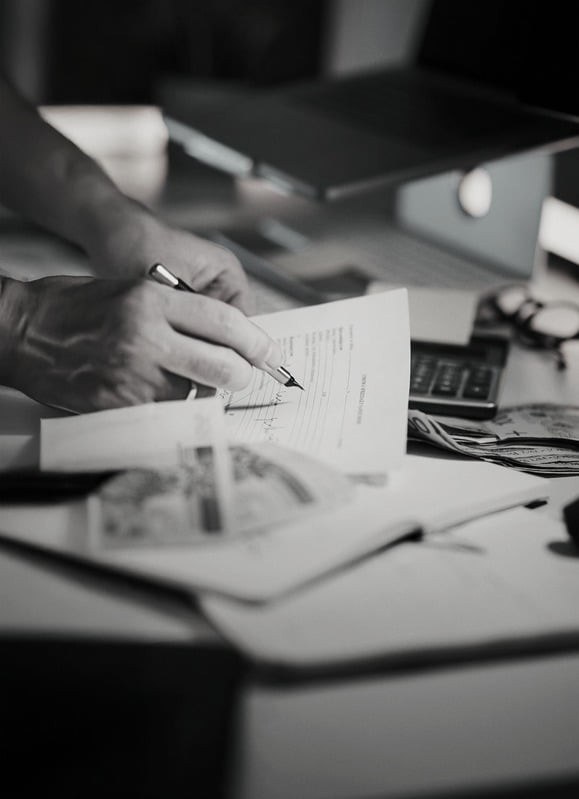 Black and white photograph of a hand holding a pen above printed documents.