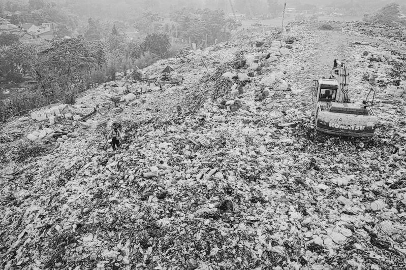 Black and white photograph of a landfill site with a tracked excavator working across mounded refuse.